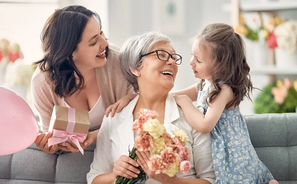 Happy mother's day! Child daughter is congratulating mom and granny giving them flowers and gift. Grandma, mum and girl smiling and hugging. Family holiday and togetherness.